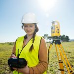 Een landmeter met een witte helm en een gele veiligheidshes voert metingen uit op een open veld. Ze houdt een controller vast en werkt met een Topcon total station, dat op een statief staat opgesteld in de achtergrond.